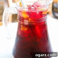 A clear glass pitcher filled with classic Spanish style sangria with ice, chunks of fruit, and a stirrer.