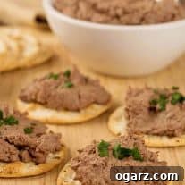 Four water crackers topped with chicken liver paté and a sprinkling of chives on a wooden board, more paté and plain crackers in the background.