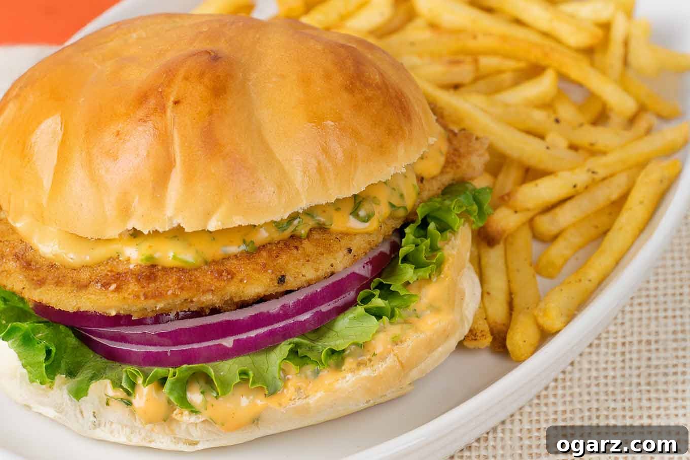 Crispy chicken sandwich on a plate with spicy sriracha mayo, fresh lettuce, and thin red onion slices, accompanied by golden oven fries for a complete meal.