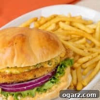 A plate with a breaded chicken sandwich topped with lettuce, red onion, and sriracha mayo on a soft roll, fries on the side.