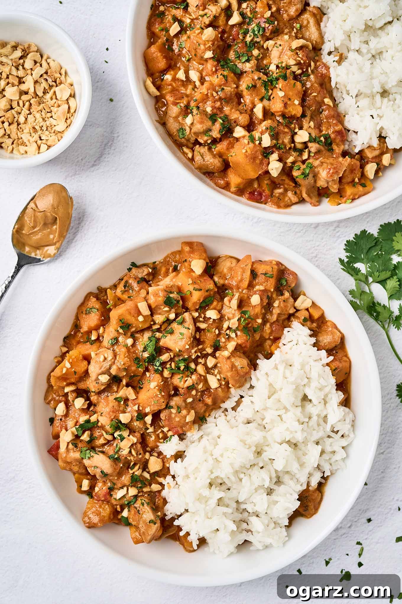 An overhead view of two bowls of West African chicken peanut stew with white rice, topped with chopped peanuts and cilantro; a bowl of extra peanuts, a spoon of peanut butter, and a sprig of cilantro to the side.
