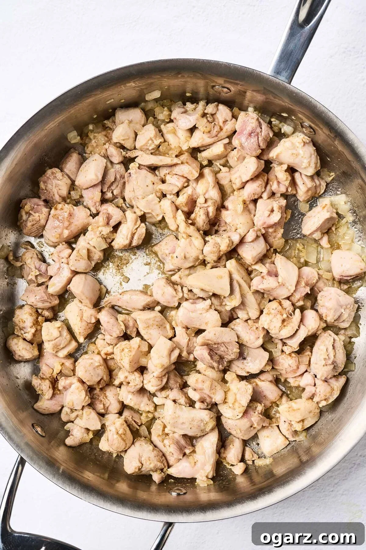 Bite sized chunks of boneless chicken thighs cooking in a sauté pan with chopped onion, garlic, and ginger.