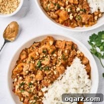 An overhead view of two bowls of West African chicken peanut stew with white rice, topped with chopped peanuts and cilantro; a bowl of extra peanuts, a spoon of peanut butter, and a sprig of cilantro to the side.
