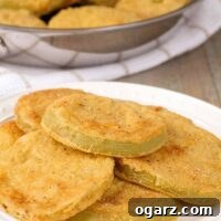 Crispy, cornmeal crusted, fried green tomatoes on a serving plate, a pan with more in the background.