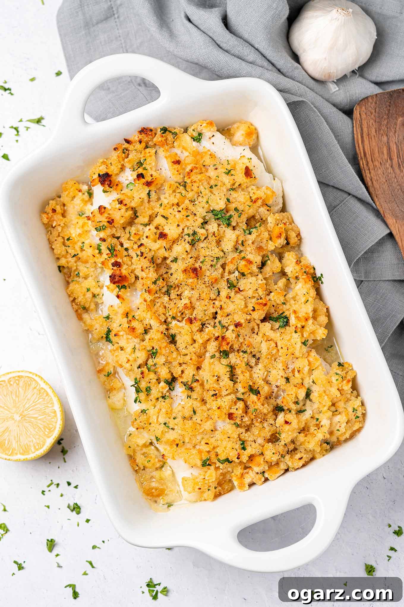 Crispy Crumb Cod Bake 2 A casserole filled with baked cod fillets topped with breadcrumb stuffing, surrounded by linen napkin, wooden server, and lemon wedge. The ultimate comfort food with a gourmet touch.