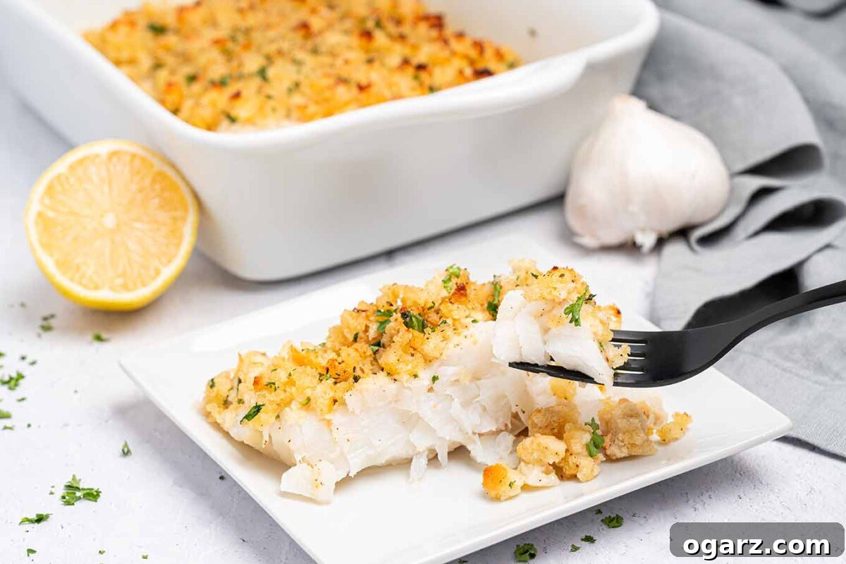 Crispy Crumb Cod Bake 7 A small plate with a single serving of baked cod with garlic breadcrumbs and black fork, casserole dish, half a lemon, and a bulb of garlic in the background, showcasing its delicious appeal.
