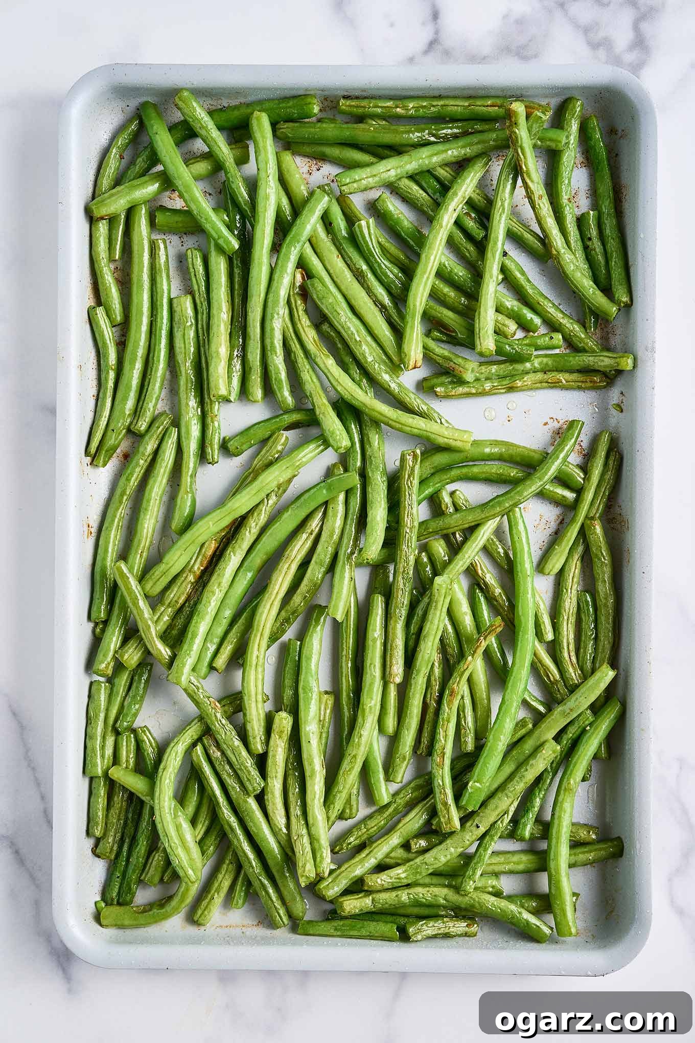 Freshly trimmed green beans, lightly coated in oil and seasoning, are spread evenly on a baking sheet, ready for oven roasting to achieve tender-crisp perfection.