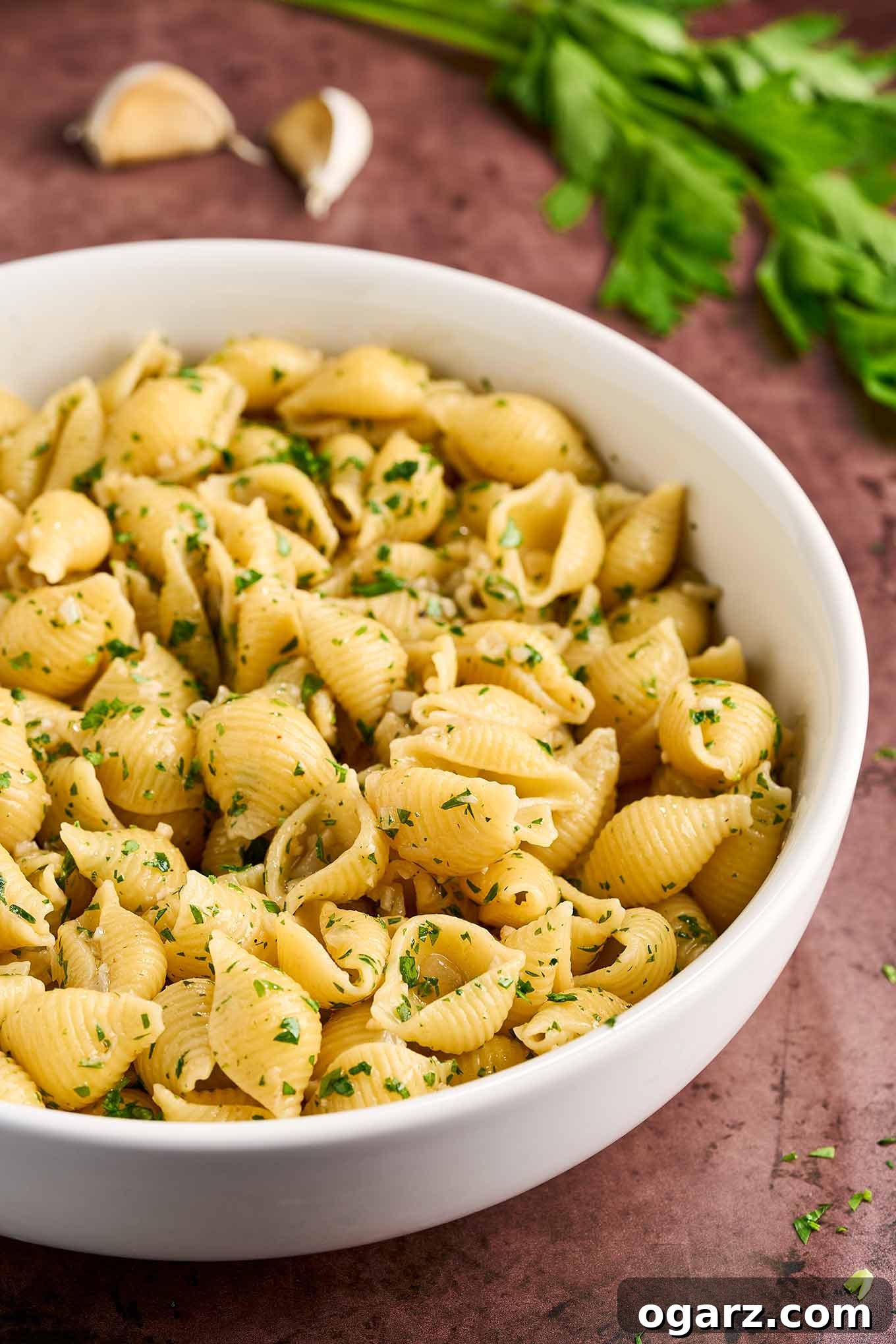 Steaming hot, medium pasta shells coated in a rich garlic butter sauce and sprinkled with vibrant green parsley, served in a white bowl.