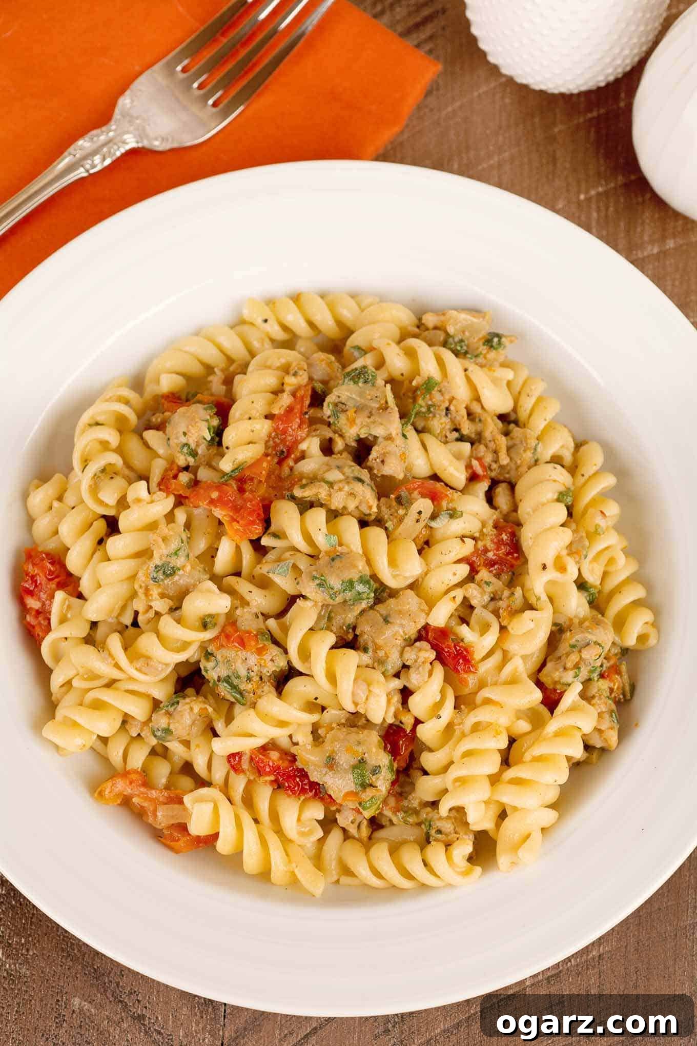A vibrant bowl of rotini pasta generously tossed with crumbled, savory Italian sausage, tender sun-dried tomatoes, and bright orange zest. A fork rests alongside, inviting a taste, complemented by a rustic linen napkin and classic salt and pepper shakers in the soft-focused background. This dish promises a unique blend of flavors, perfect for a satisfying meal.