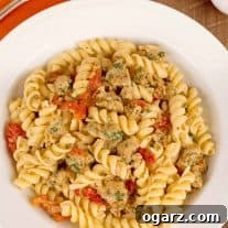 A vibrant bowl of rotini pasta generously tossed with crumbled, savory Italian sausage, tender sun-dried tomatoes, and bright orange zest.