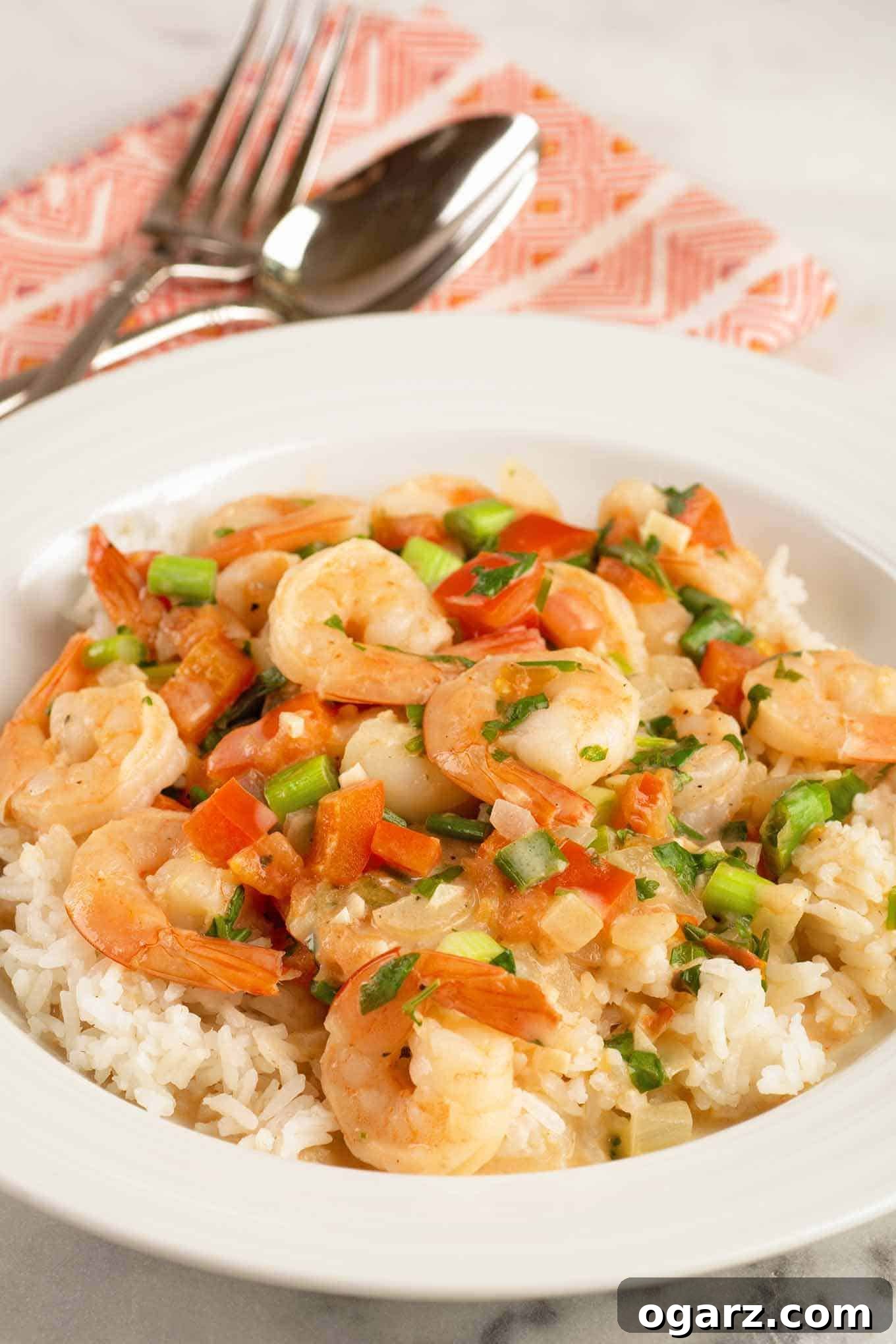Brazilian Coconut Shrimp Stew 2 A shallow bowl of steamed rice topped with vibrant Brazilian shrimp stew in creamy coconut milk, garnished with red pepper, tomato, scallions, cilantro, and a lime wedge. A fork and spoon rest on a napkin in the background, inviting a delicious meal.