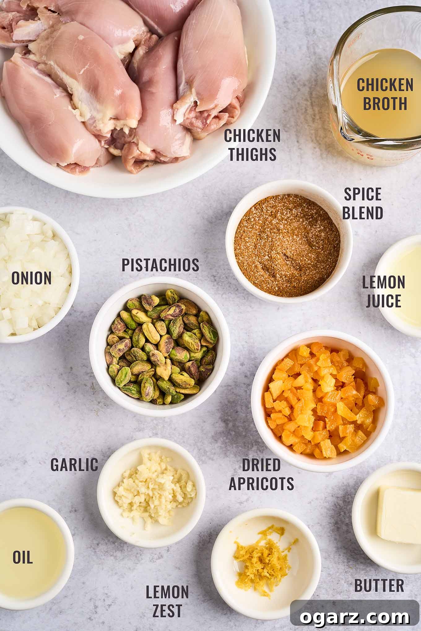 A labeled, overhead view of the ingredients for Persian apricot chicken; boneless, skinless chicken thighs, chicken broth, spice blend, lemon juice and zest, dried apricots, butter, garlic, oil, and chopped onion.