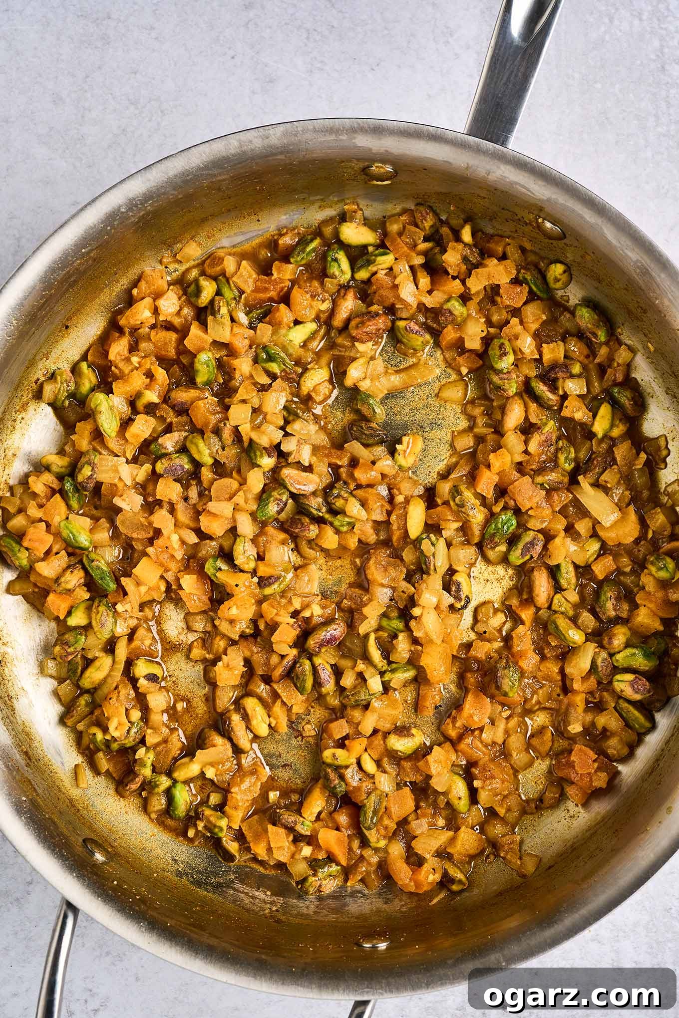 A frying pan with sautéed onion and garlic, diced dried apricots, and pistachio nuts seasoned with a blend of salt, sugar, turmeric, coriander, cumin, cinnamon, cayenne, and black pepper.