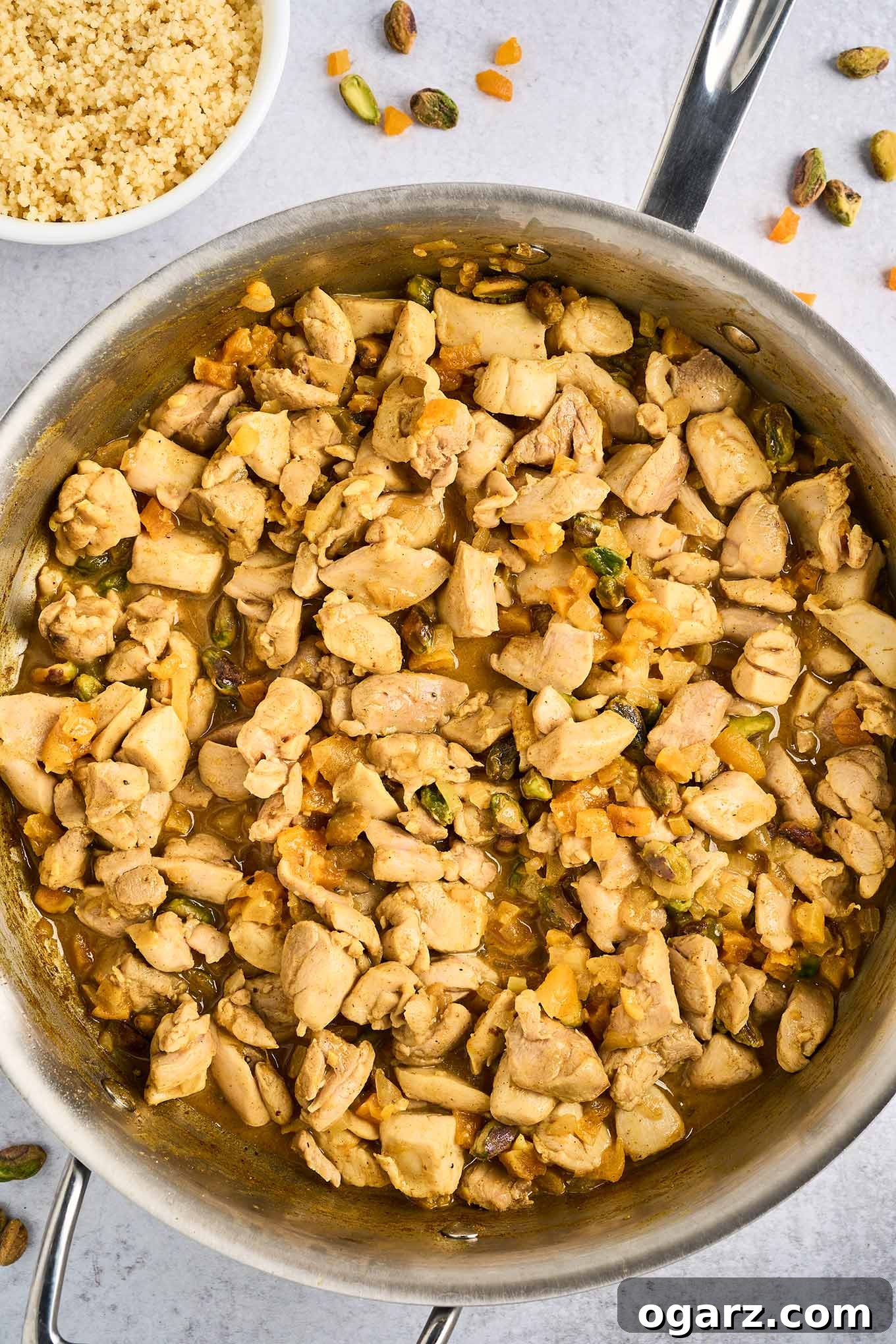 A frying pan filled with chunks of tender chicken thighs combined with spices, dried apricots, pistachio nuts, sautéed garlic and onion, lemon juice and zest, and butter, a bowl of couscous in the background.