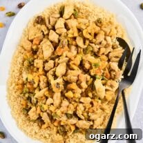 An oval serving platter of couscous topped with Persian style apricot chicken and pistachios with serving utensils, surrounded by a prep bowl of the spice blend, dried apricots, and chopped pistachios.