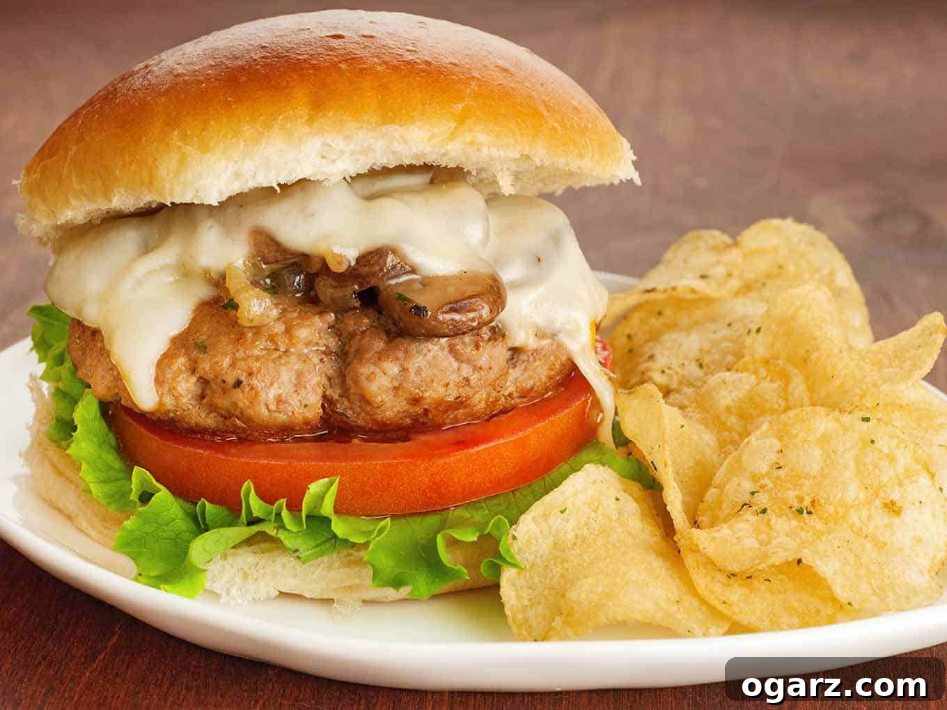 Umami Turkey Mushroom Burgers 2 A perfectly pan-seared and braised turkey burger, richly topped with sautéed golden mushrooms and caramelized onions, and draped with melted muenster cheese. It's nestled in a soft bun with crisp lettuce and a slice of ripe red tomato, served alongside a scattering of crunchy potato chips.