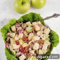 A lettuce lined bowl of Waldorf salad made with crisp apples, fresh orange zest, dried cranberries and toasted pecans, two Granny Smith apples in the background.
