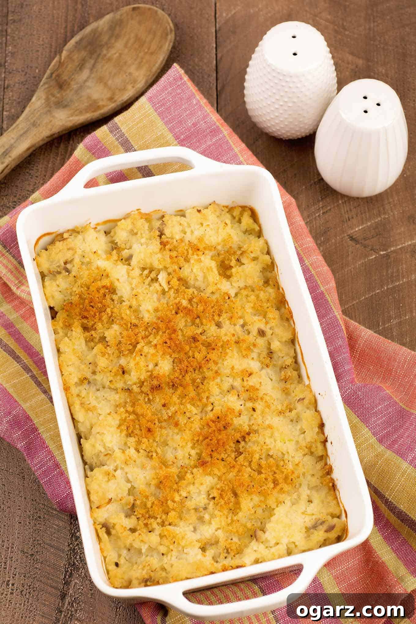 Cheesy Cauliflower Rice Gratin 2 A white, oblong casserole dish of crumb-topped, riced cauliflower gratin on a tea towel with white salt and pepper shakers in the background.
