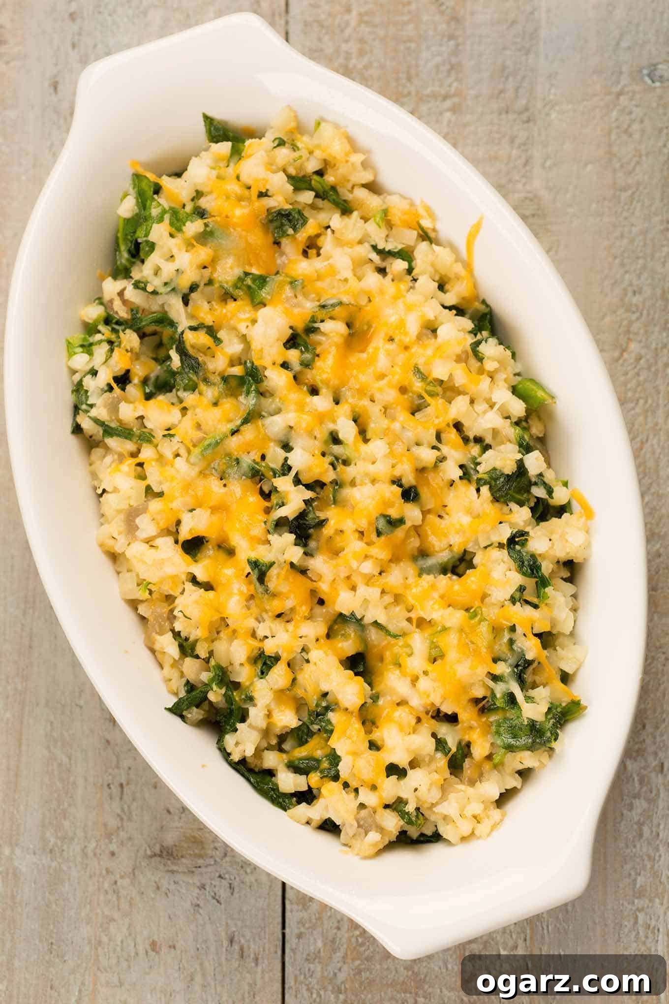 Cheesy Cauliflower Rice Gratin 3 Overhead view of a creamy riced cauliflower gratin with melted cheese and spinach in a white oval dish.