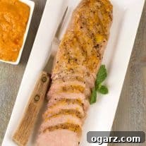 An oblong platter with a partially carved, grilled pork tenderloin topped with peach barbecue sauce, serving fork and extra sauce on the side.