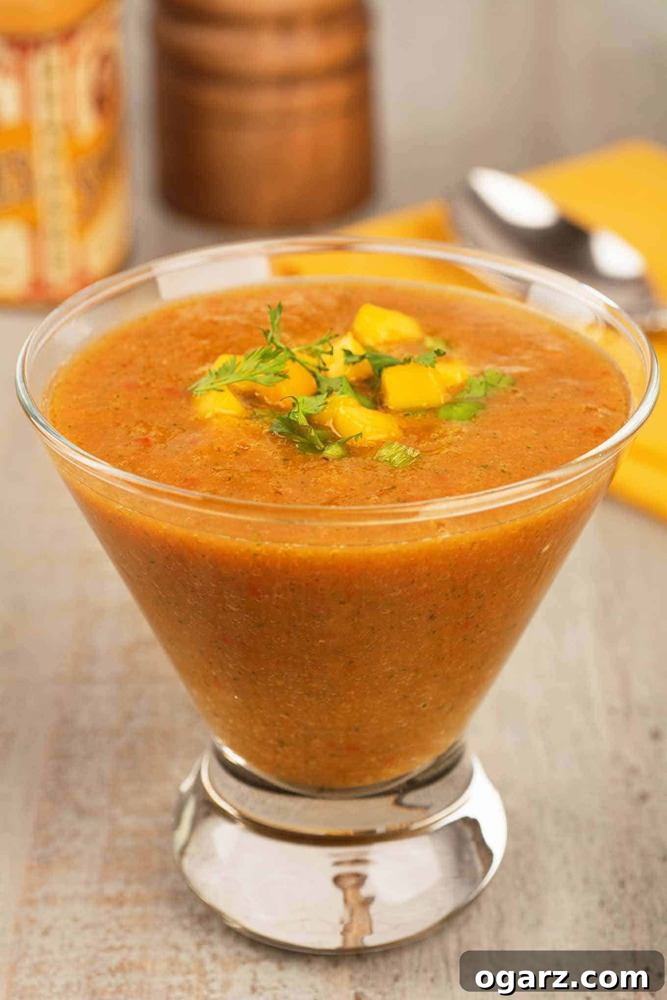 A vibrant serving of mango gazpacho soup, garnished with fresh mango cubes and cilantro leaves, presented in a clear glass. A spoon, napkin, pepper mill, and spice jar are softly blurred in the background, suggesting a gourmet dining experience.