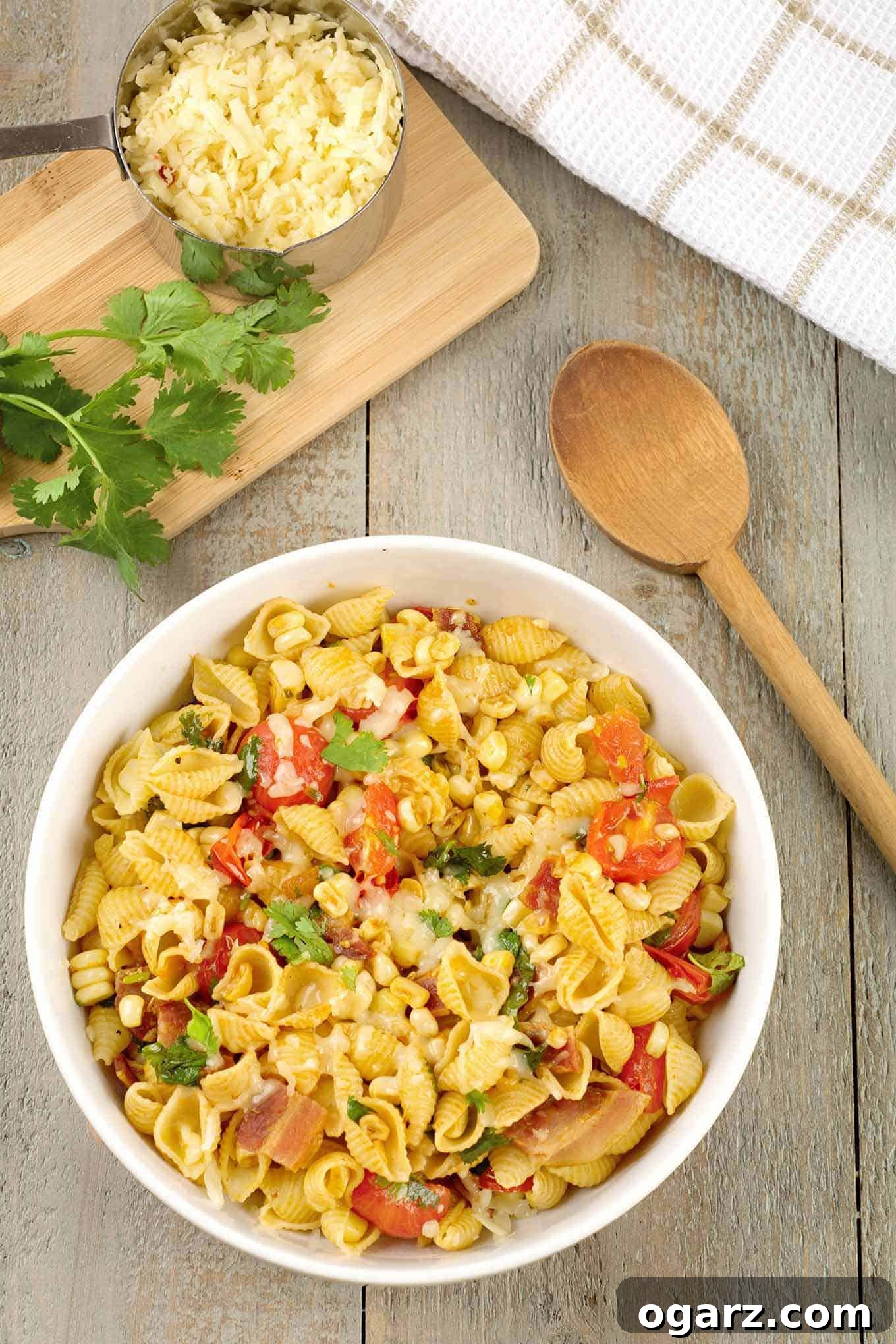 A serving bowl of pasta shells with bacon, corn, tomatoes, chipotle, cheese, and Southwest seasonings, with a cutting board, cilantro, and spoon to the side.