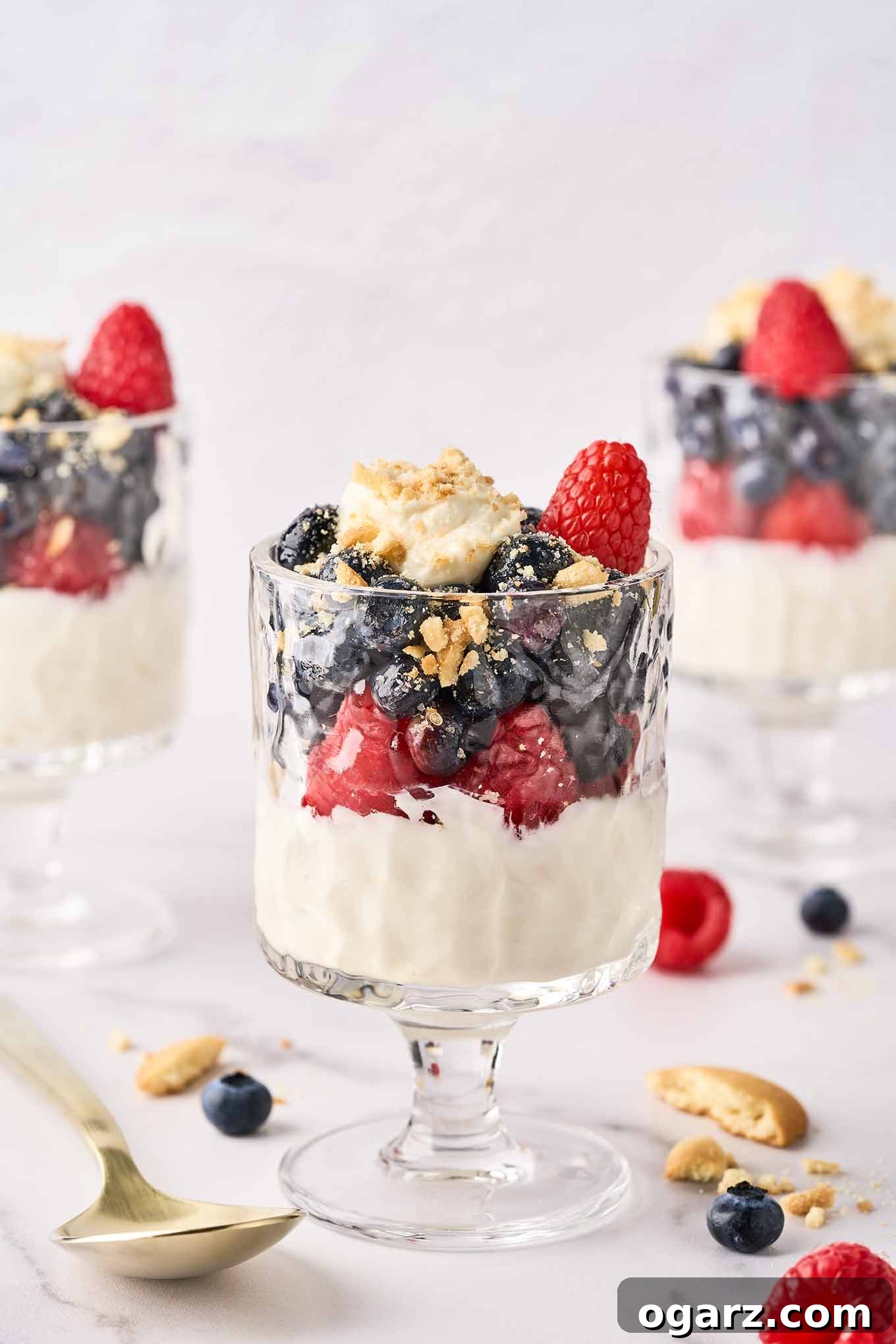 Three elegant no-bake cheesecake parfaits, each generously topped with vibrant blueberries, fresh raspberries, and delicate crumbled vanilla cookies. They are presented in clear, footed parfait glasses, artfully arranged on a rustic wooden surface amidst scattered cookie crumbs, whole fresh berries, and a vintage silver spoon, inviting immediate indulgence.