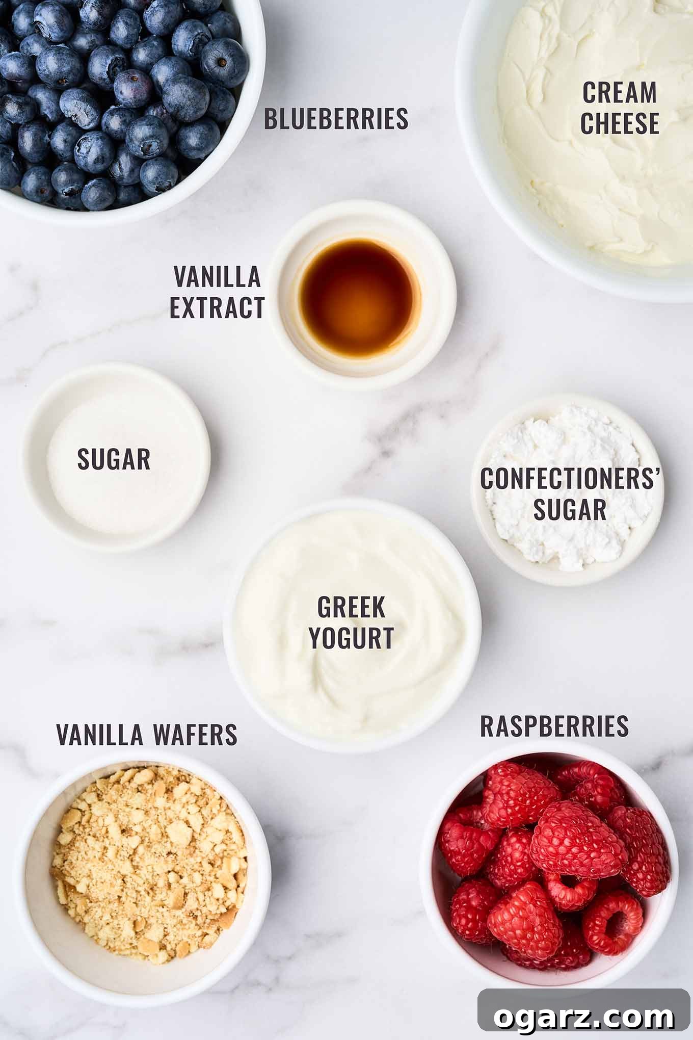 An inviting overhead shot showcasing all the fresh ingredients needed for no-bake cheesecake parfaits with a colorful array of berries. The display includes a block of softened cream cheese, a container of creamy Greek vanilla yogurt, a small bowl of confectioners' sugar, a stack of golden vanilla wafers, vibrant fresh raspberries and blueberries, and a bottle of pure vanilla extract, all artfully arranged on a light background.