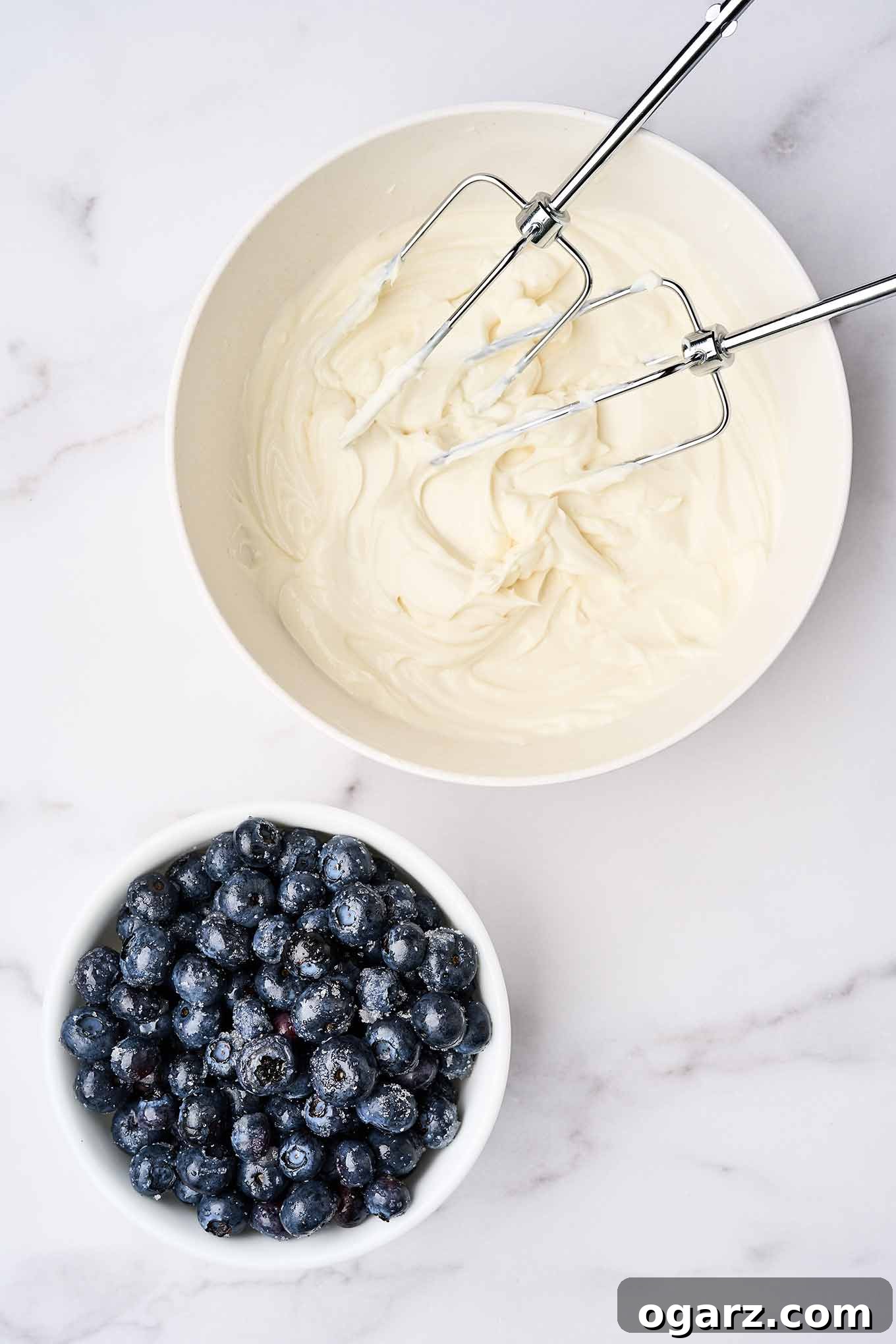A vibrant scene of cheesecake parfait preparation, highlighting the smooth, whipped cream cheese and Greek vanilla yogurt mixture ready in a large mixing bowl. An electric mixer with beaters rests beside the bowl, while a separate small bowl holds sugared blueberries, glistening and ready for assembly. This image captures the essence of fresh, simple ingredients coming together for a delicious dessert.