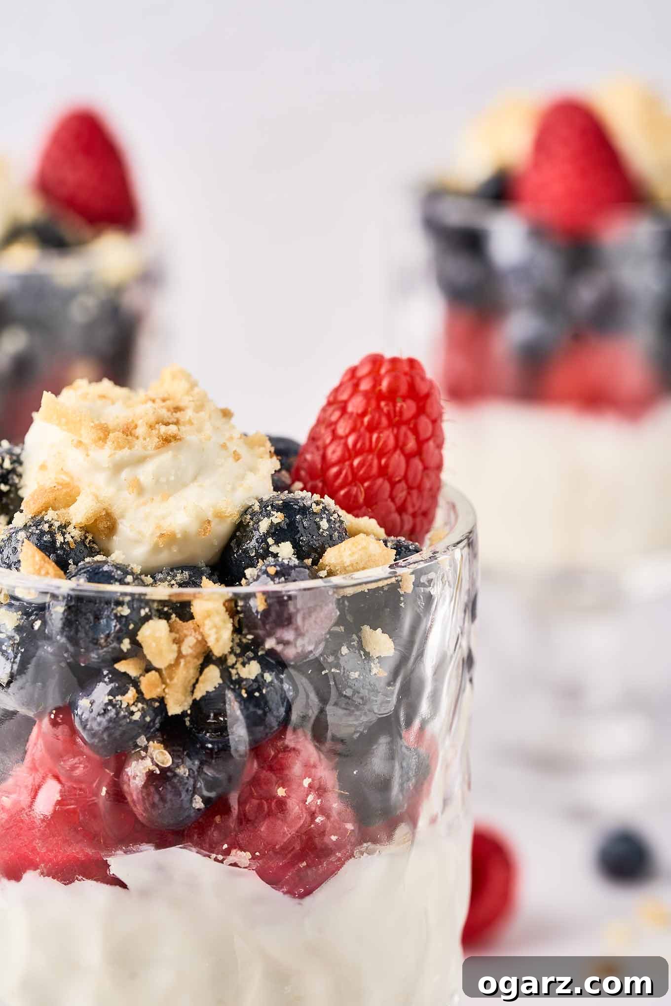 A close-up, mouth-watering view of a single parfait glass, brimming with a layered no-bake cheesecake. The creamy white cheesecake base is beautifully topped with a vibrant medley of fresh raspberries, glistening sugared blueberries, and a scattering of golden crumbled vanilla wafers, showcasing the irresistible textures and colors of this delightful dessert.