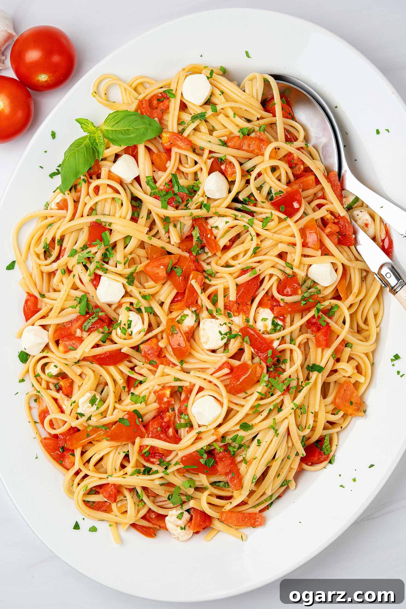 Vibrant Caprese Pasta 2 Caprese pasta on a platter with serving utensil and fresh basil garnish, surrounded by whole tomatoes and garlic cloves.