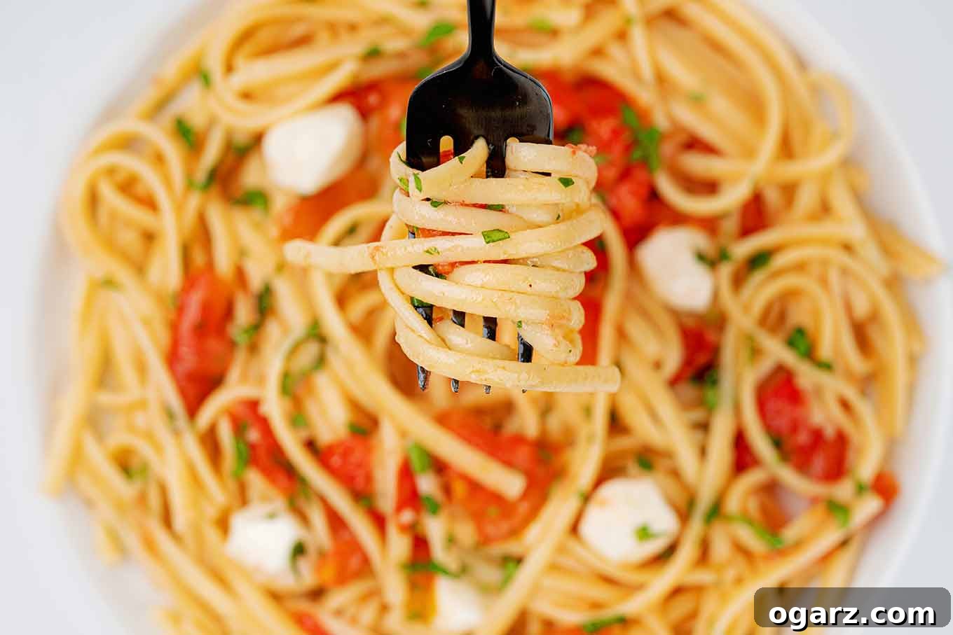 Vibrant Caprese Pasta 5 A closeup view of linguine twirled around a black fork and held above a serving of caprese pasta.