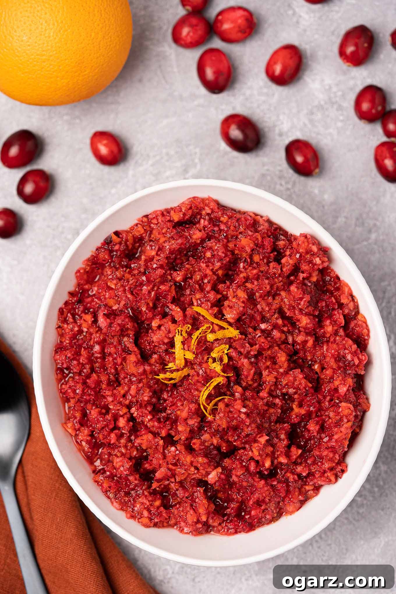 Effortless Cranberry Orange Zing 2 A serving bowl of fresh cranberry orange relish garnished with orange zest, surrounded by scattered cranberries, an orange, and a serving spoon.