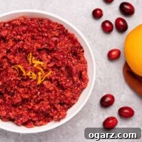 Effortless Cranberry Orange Zing 5 A serving bowl of fresh cranberry orange relish garnished with orange zest, on a gray background with scattered cranberries to the right.
