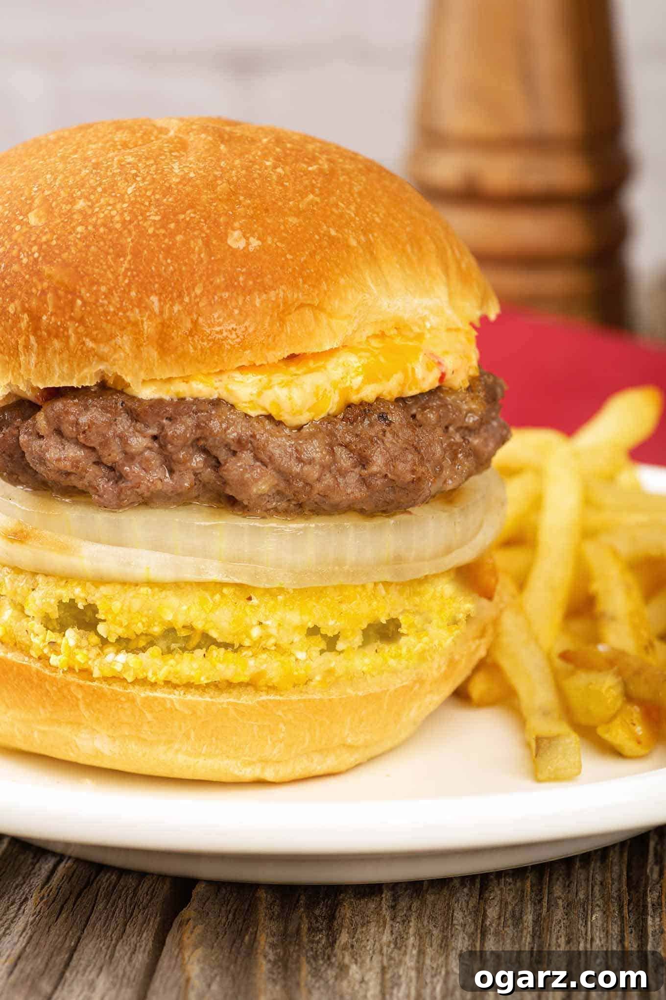 Southern Fried Green Tomato Burgers 2 A close view of a grilled burger stacked on a bun with slices of fried green tomato and sweet onion, topped with pimento cheese, with fries on the side.