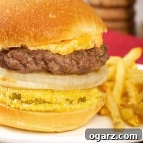 A close view of a grilled burger stacked on a bun with slices of fried green tomato and sweet onion, and a topping of pimento cheese - fries on the side.