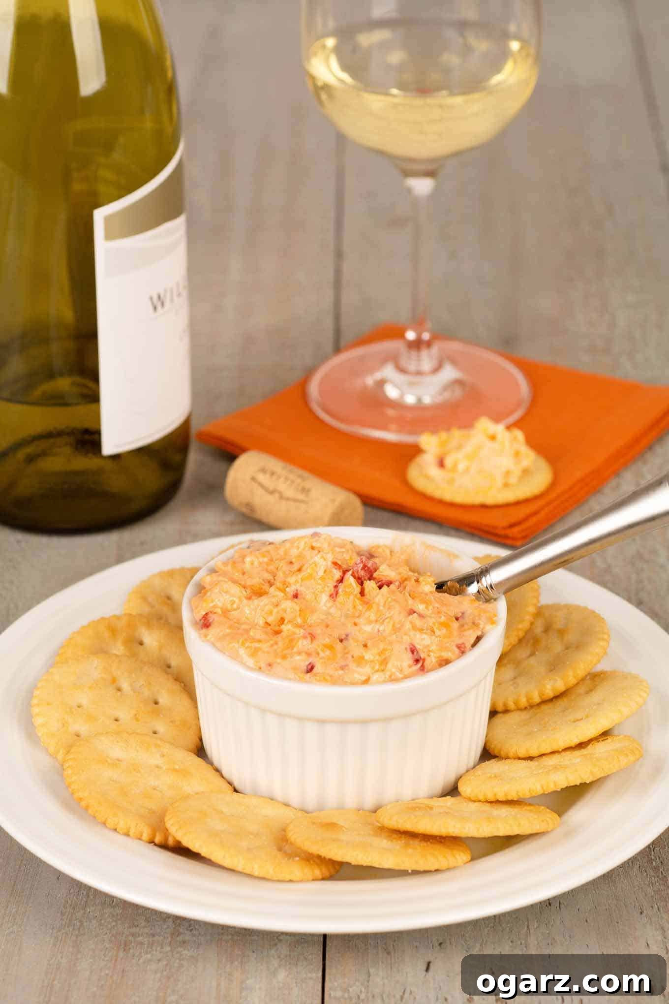 Southern Pimento Cheese 2 A white crock filled with vibrant pimento cheese, surrounded by crispy crackers on a rustic wooden plate. In the blurred background, a wine bottle, a wine glass, and a cracker topped with the cheese suggest a perfect pairing.
