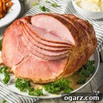 A large, spiral-sliced, glazed ham on a serving platter, garnished with fresh parsley and accompanied by mashed potatoes in the background.