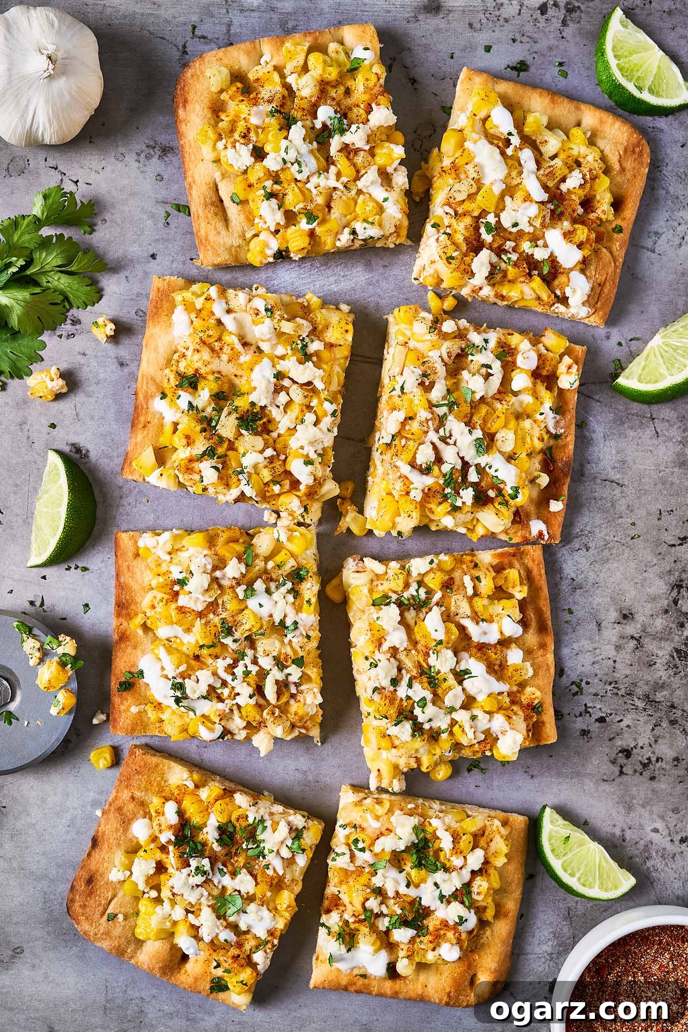 Mexican street corn flatbread cut into pieces on a baking sheet and garnished with fresh cilantro; lime wedges, garlic, and elote seasoning to the side.