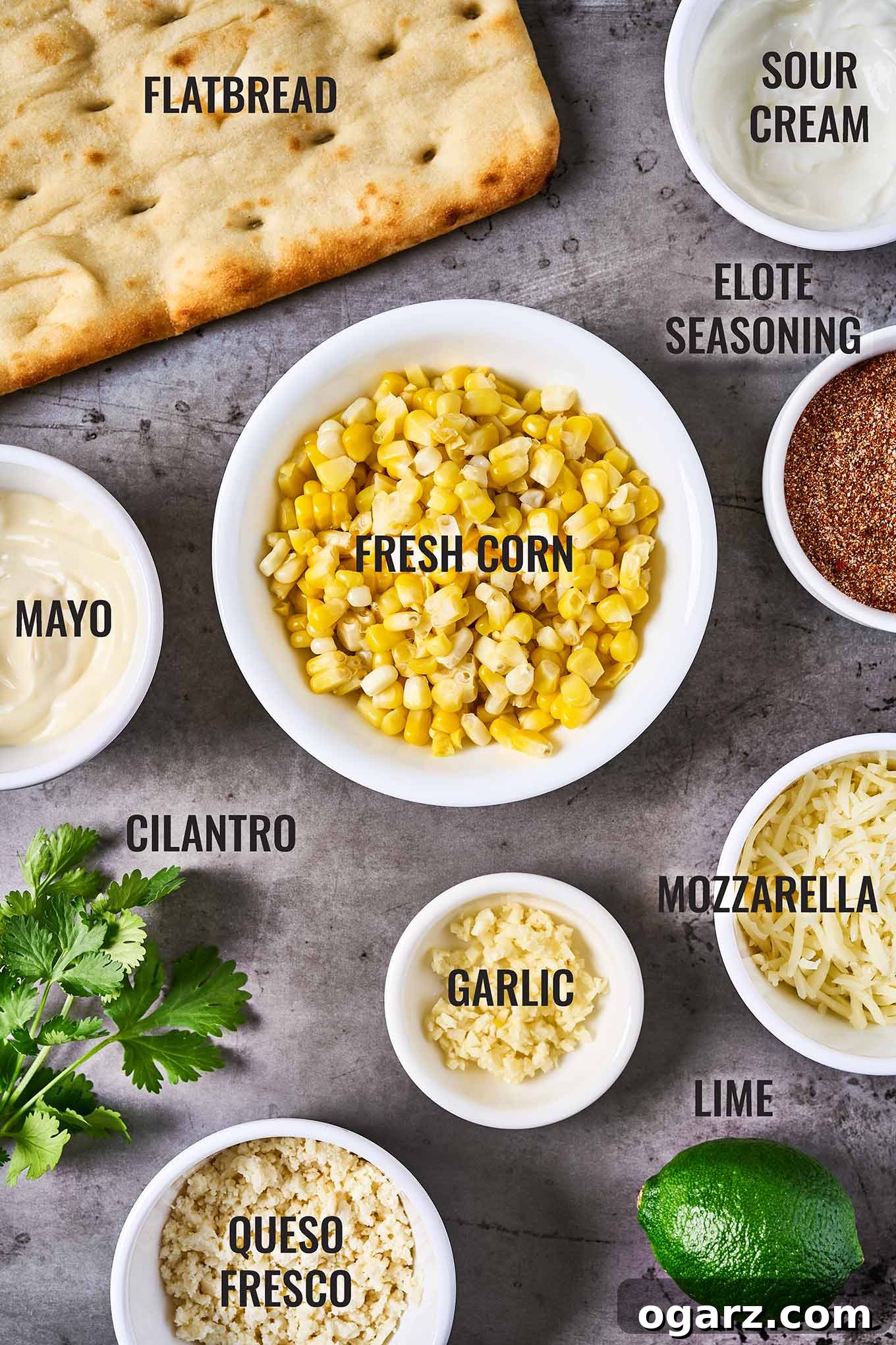 A labeled, overhead view of the ingredients for Mexican street corn flatbread including flatbread, corn kernels, elote seasoning, queso fresco, lime, and cilantro.