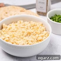 Fresh, homemade breadcrumbs in a bowl with a slice of bread, a prep bowl of chopped parsley, a bread knife, and some seasoning in the background.