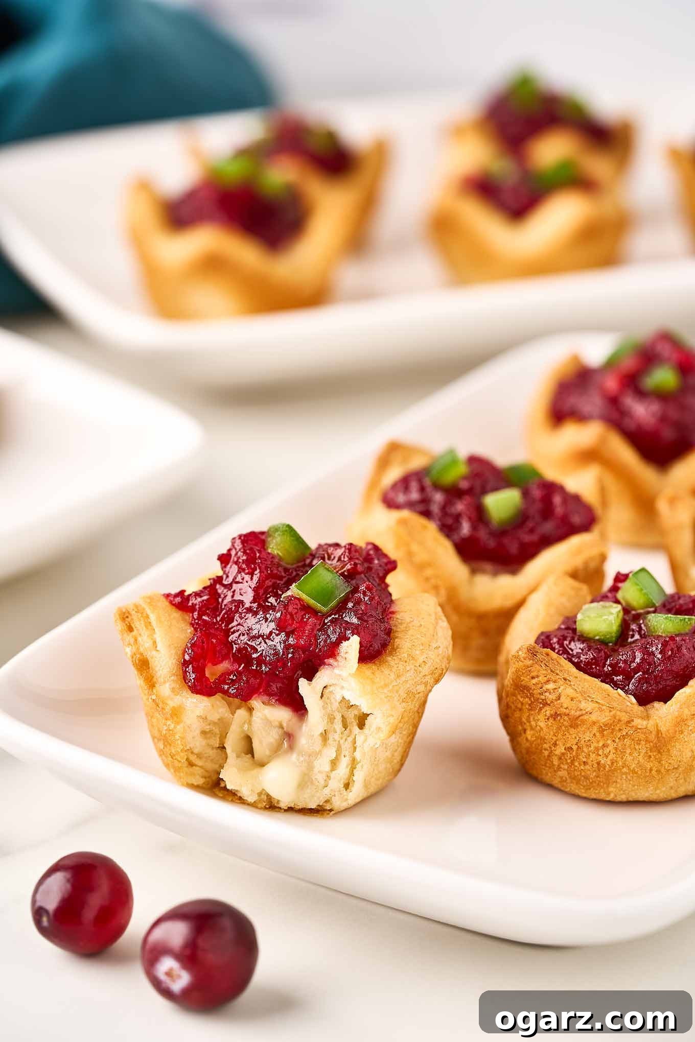 A close up of cranberry brie bites topped with diced jalapeno pepper, one with a bite out it showing the melted brie and cranberry jam topping.