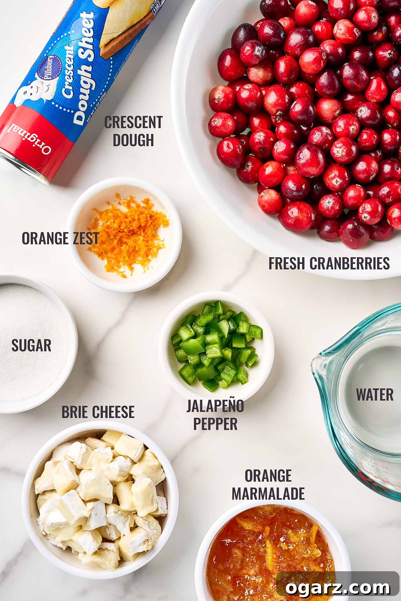A labeled, overhead view of the ingredients for cranberry brie bites including cranberries, orange zest, marmalade, jalapeno pepper, brie cheese, and crescent dough.