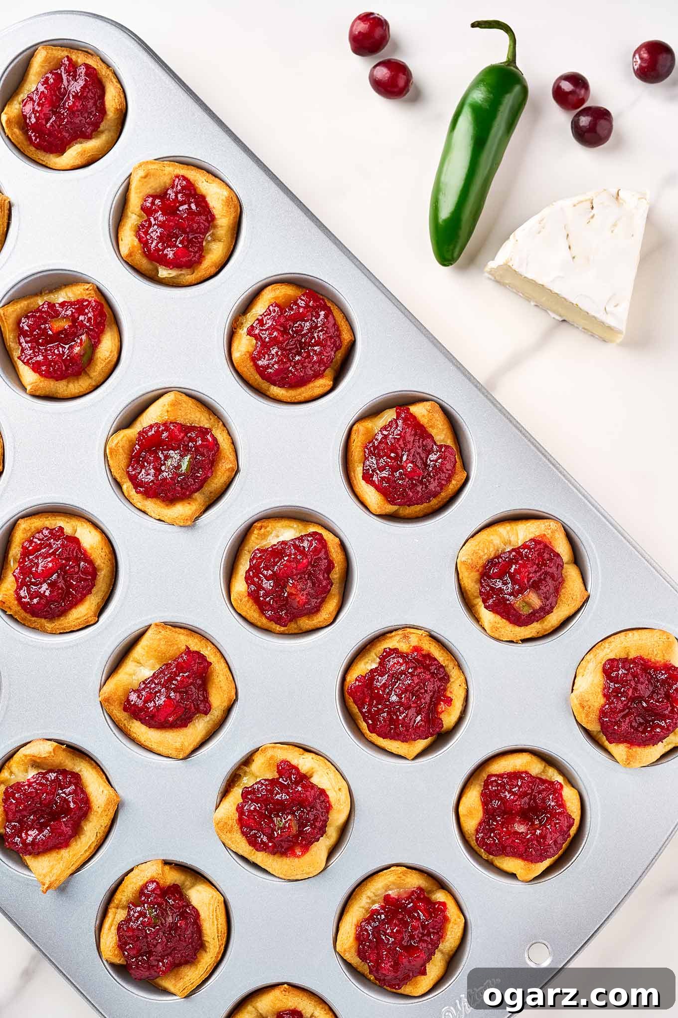 A mini muffin tin of brie bites just topped with homemade cranberry jam, scattered cranberries, a wedge of brie, and a jalapeno pepper to the side.