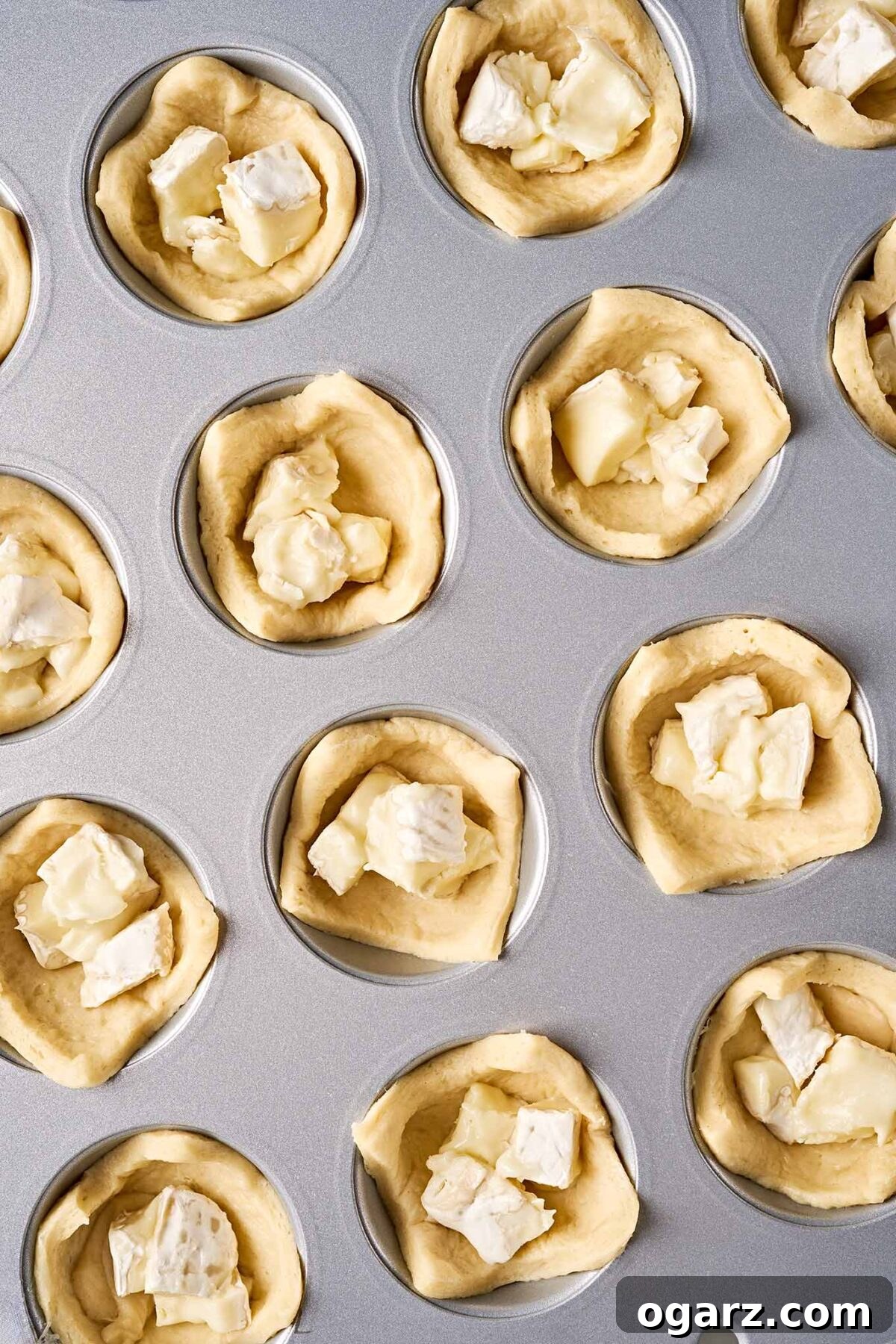 Squares of crescent dough filled with chunks of brie in a mini muffin tin, ready to be baked.