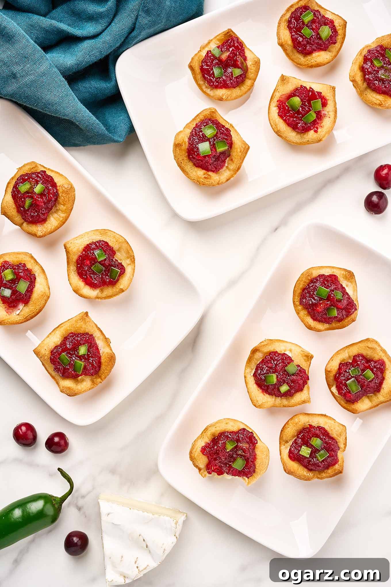Three white appetizer plates with jalapeno and cranberry brie bites on them, on a marble surface, a blue linen napkin, wedge of brie, and cranberries to the side.