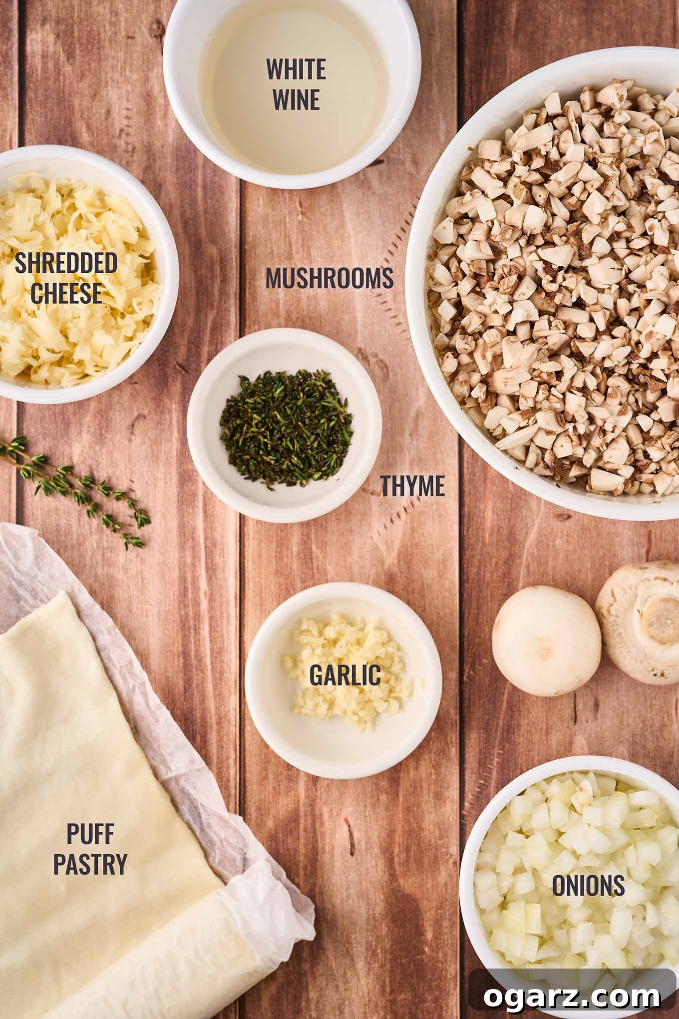 Umami Mushroom Puffs 3 A labeled, overhead view of the ingredients for mushroom puffs including pastry dough, mushrooms, onions, garlic, thyme, shredded cheese, and white wine.