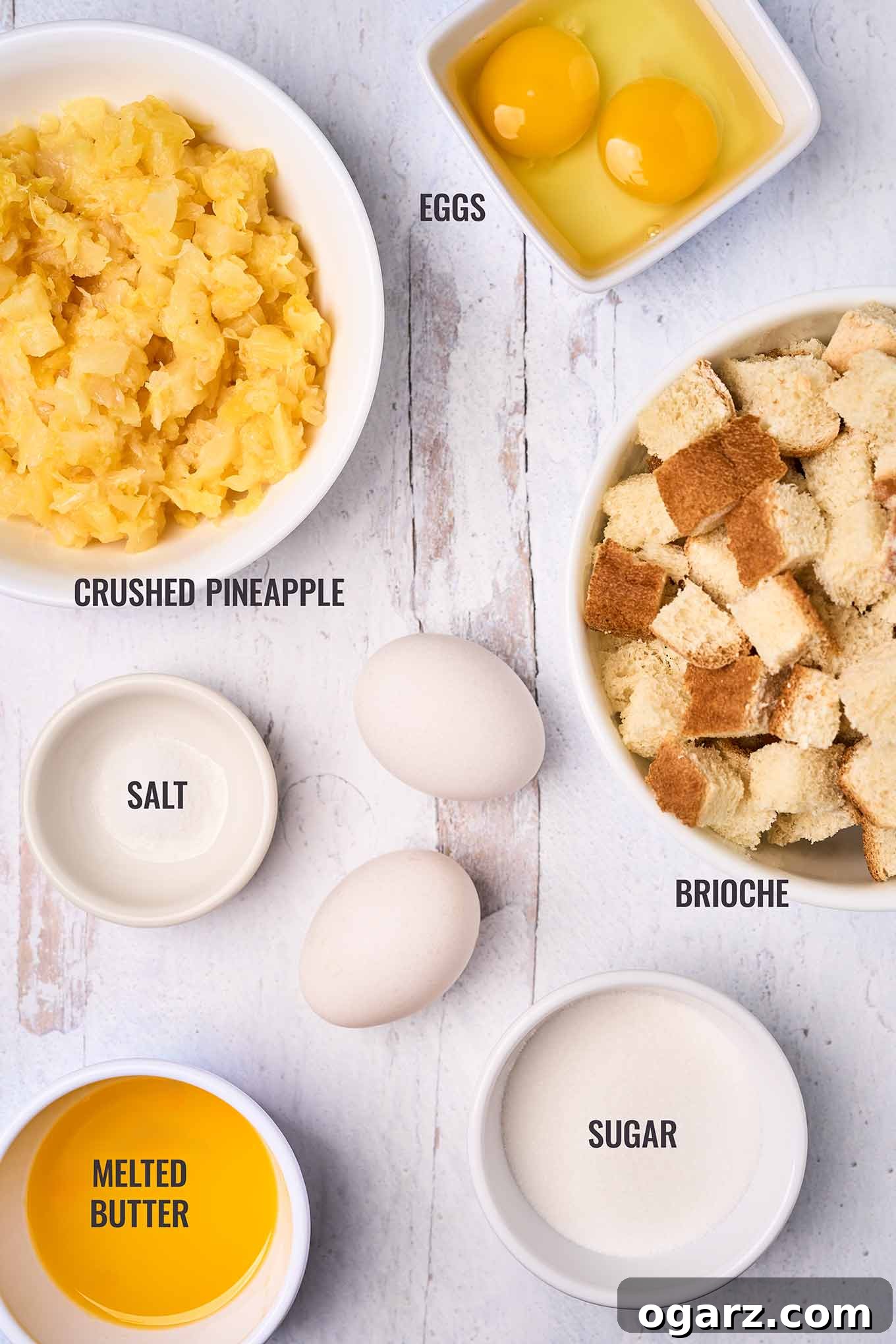 Golden Pineapple Gratin 3 A beautifully arranged overhead shot of scalloped pineapple ingredients, featuring golden brioche bread cubes, a bowl of bright crushed pineapple, fresh eggs, granulated sugar, a pinch of salt, and a small dish of melted butter.