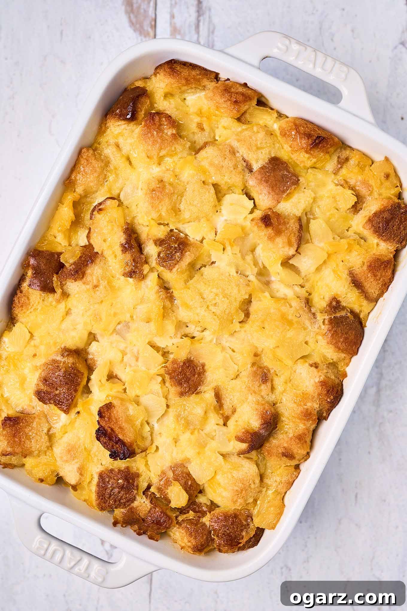 Golden Pineapple Gratin 6 A white oblong casserole dish brimming with golden-brown scalloped pineapple, artfully placed on a distressed white wooden surface, ready to be enjoyed as a comforting and delicious meal.