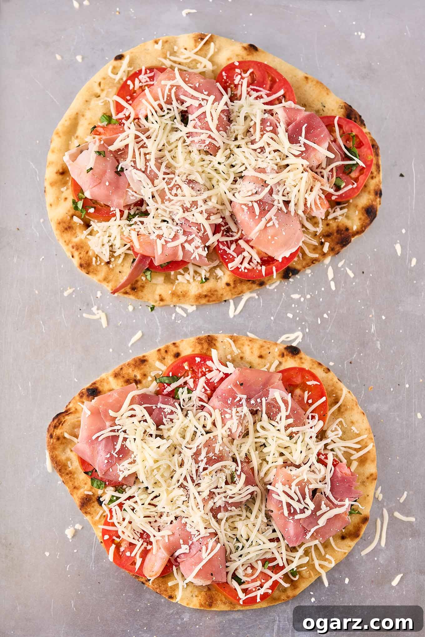 Two naan flatbread pizzas on a baking sheet, topped with prosciutto-wrapped shrimp, tomatoes, and mozzarella and ready for the oven.
